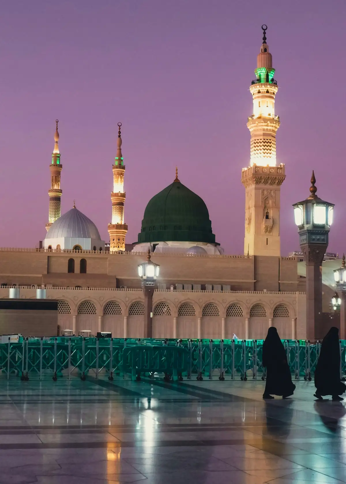 Two women in black abayas walking past the Prophet's Mosque in Madinah at dusk